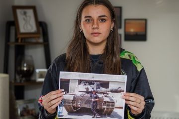 A young girl holds a photo of her dad who served in the Army. He is standing at the front of a tank